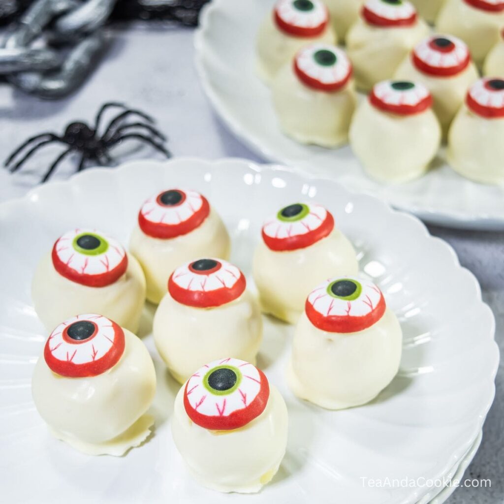 White chocolate truffles decorated with candy eyeballs are arranged on white plates, with a plastic black spider in the background.