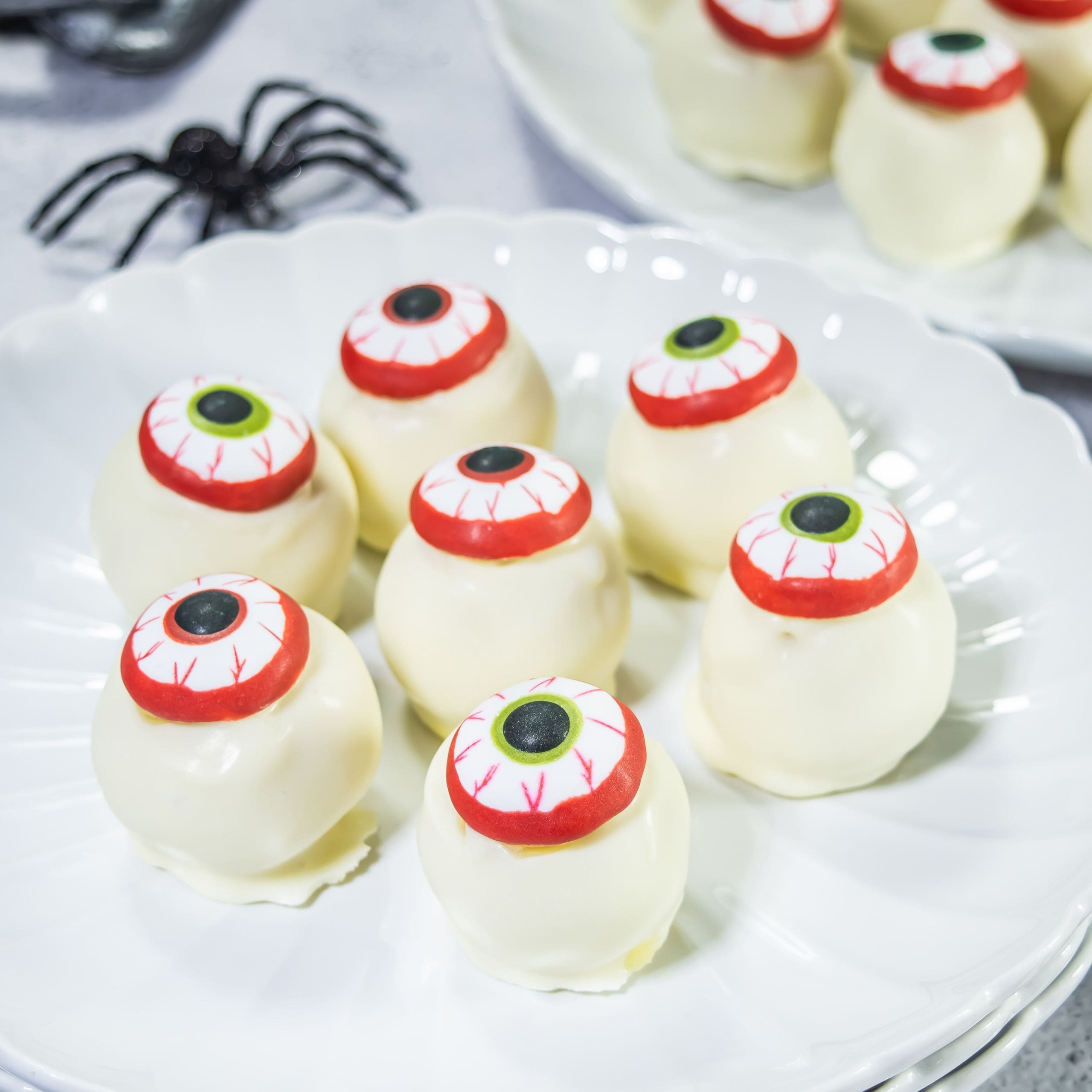 White chocolate truffles decorated with candy eyes, arranged on a white plate. A plastic black spider is visible in the background.