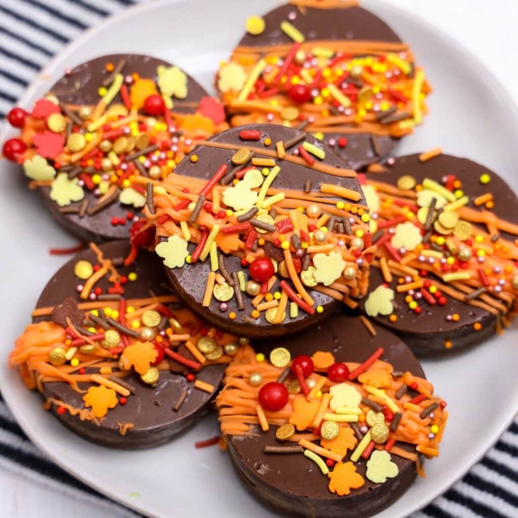 Chocolate covered Oreos for fall. A plate of chocolate-covered cookies decorated with orange, yellow, and red fall-themed sprinkles and drizzles.