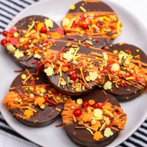 Chocolate covered Oreos for fall. A plate of chocolate-covered cookies decorated with orange, yellow, and red fall-themed sprinkles and drizzles.