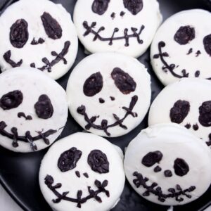 Seven round cookies with white icing are decorated with black icing to resemble Jack Skellington faces from The Nightmare Before Christmas, arranged on a black plate.