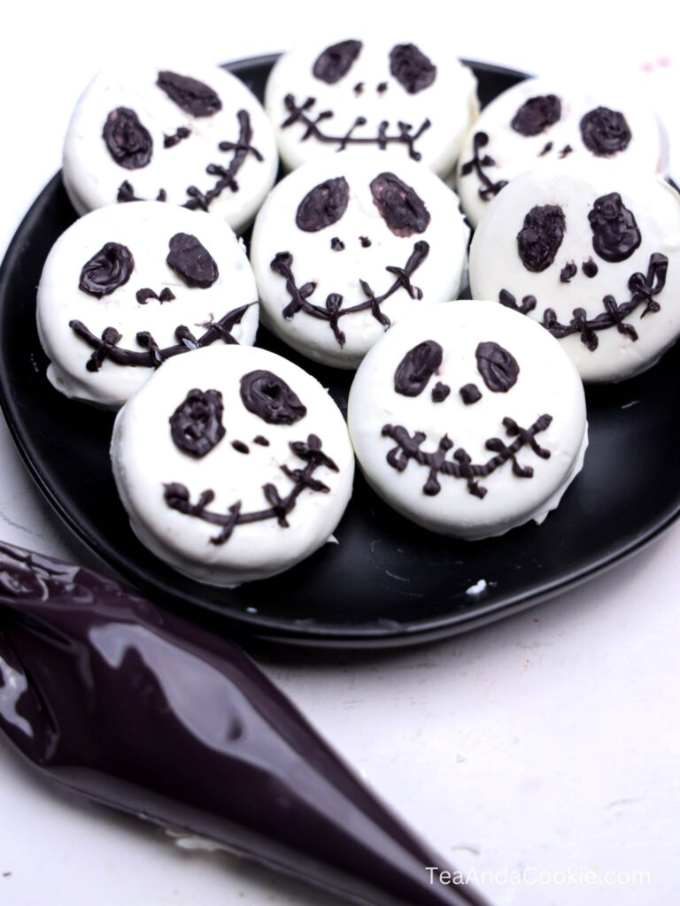 A black plate with eight white, round cookies decorated with black Jack Skellington faces. A piping bag with black icing is placed beside the plate.