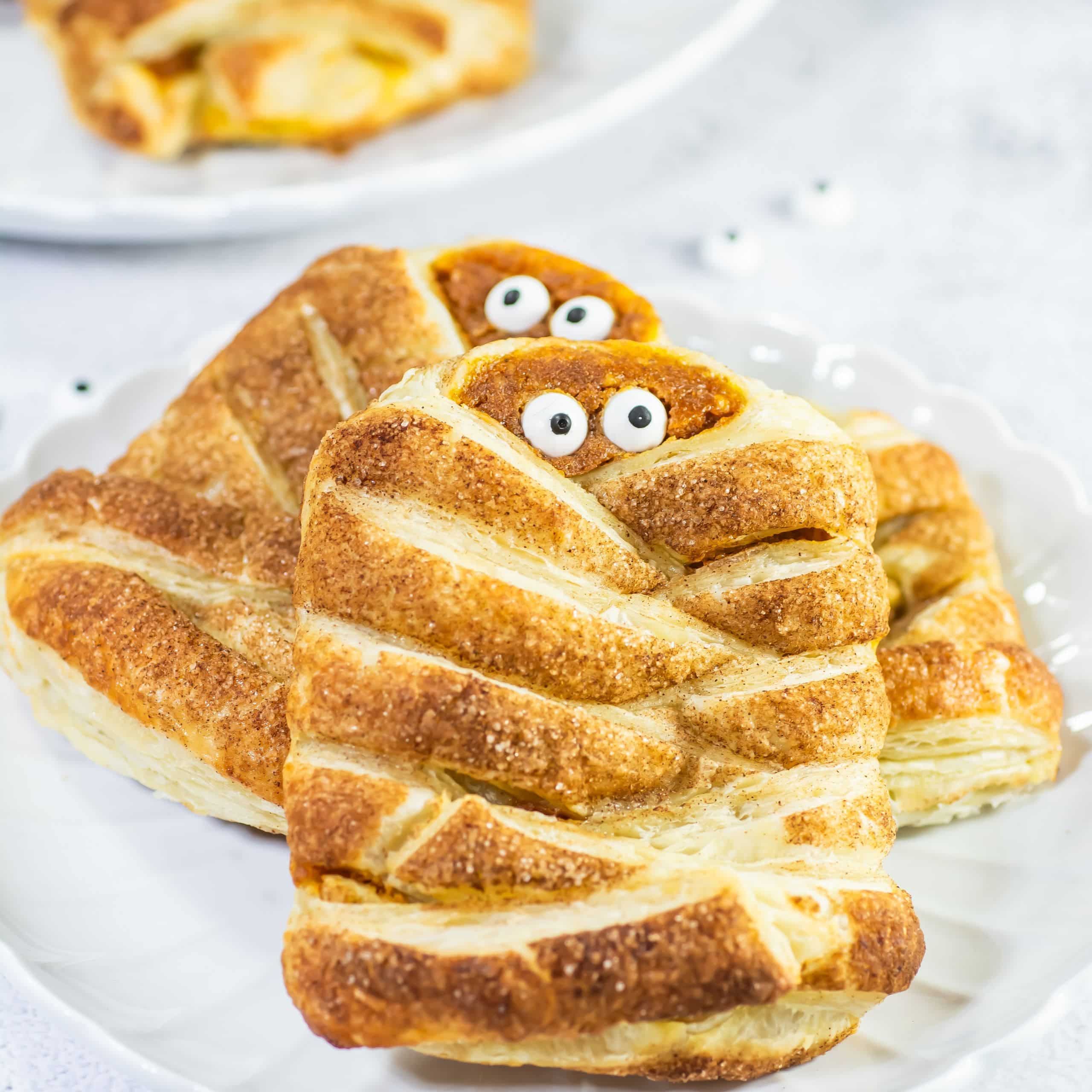 Two puff pastries decorated to look like mummies with candy eyes, arranged on a white plate.