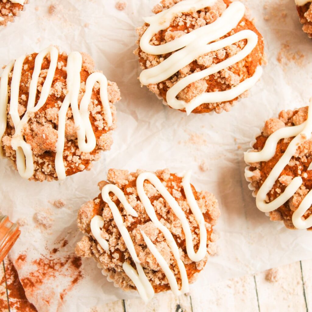 Four crumb-topped muffins with white icing drizzle are arranged on parchment paper over a rustic wooden surface.