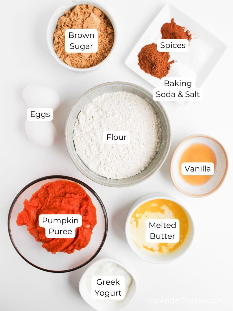 Top-down view of labeled baking ingredients in separate bowls, including flour, brown sugar, spices, baking soda, salt, eggs, pumpkin puree, melted butter, vanilla, and Greek yogurt.