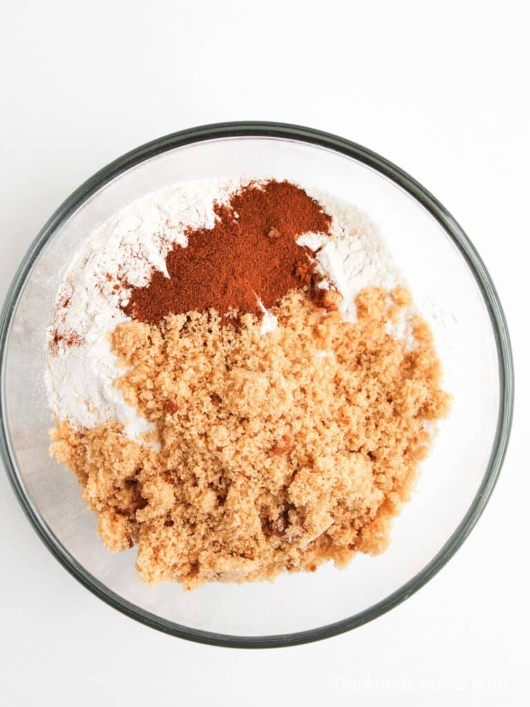 A glass bowl containing flour, brown sugar, and ground cinnamon on a white surface.