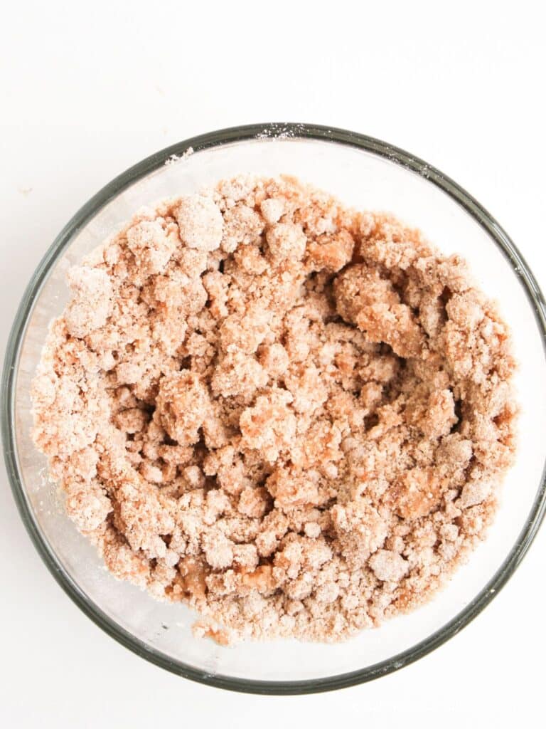 A clear glass bowl filled with a crumbly mixture of light brown sugar and flour, seen from above against a white background.