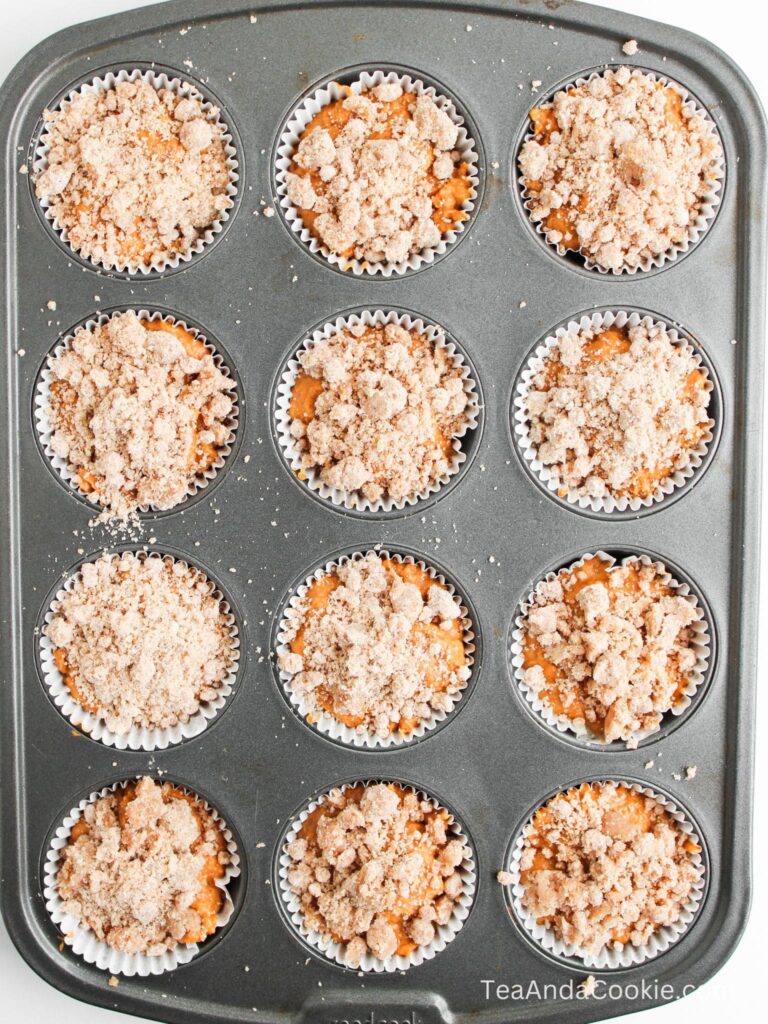 A muffin tin holds twelve unbaked muffins in paper liners, each topped with a layer of crumbly streusel.