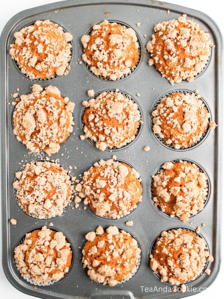 A metal muffin tray holds twelve baked muffins topped with a crumbly streusel.