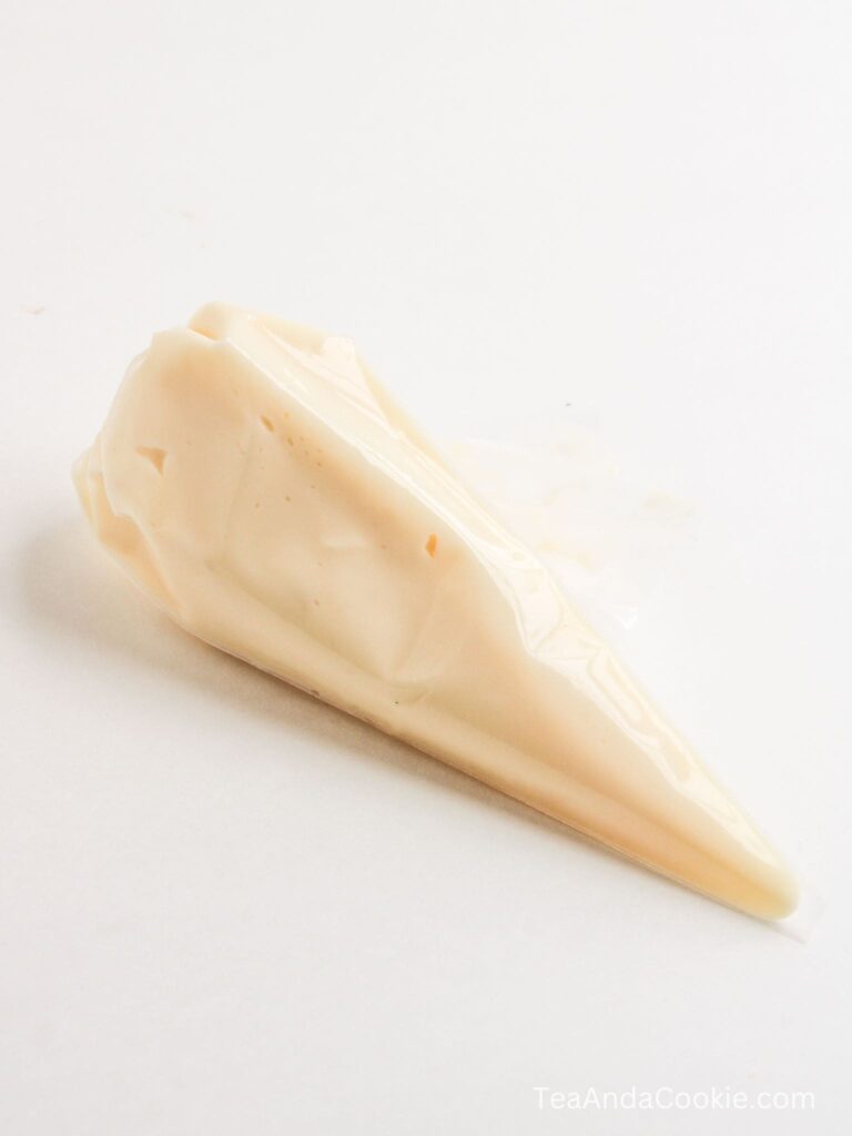 A small, clear plastic piping bag filled with light-colored icing or cream rests on a white surface.