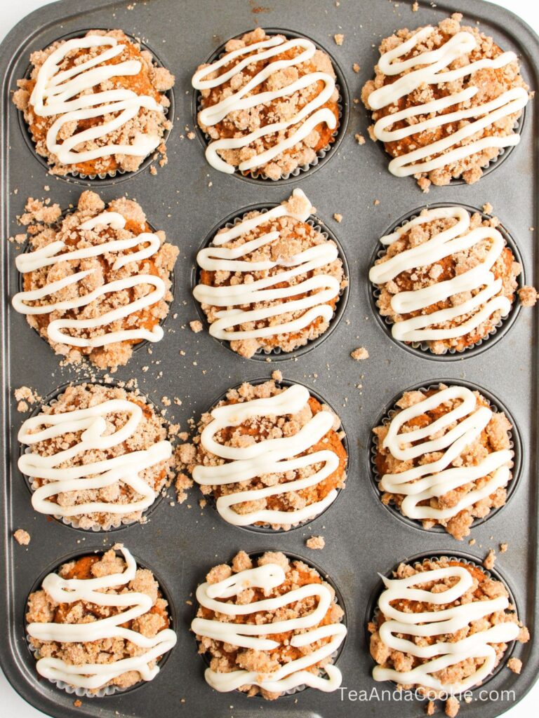 A muffin tin filled with twelve crumb-topped muffins, each drizzled with white icing.