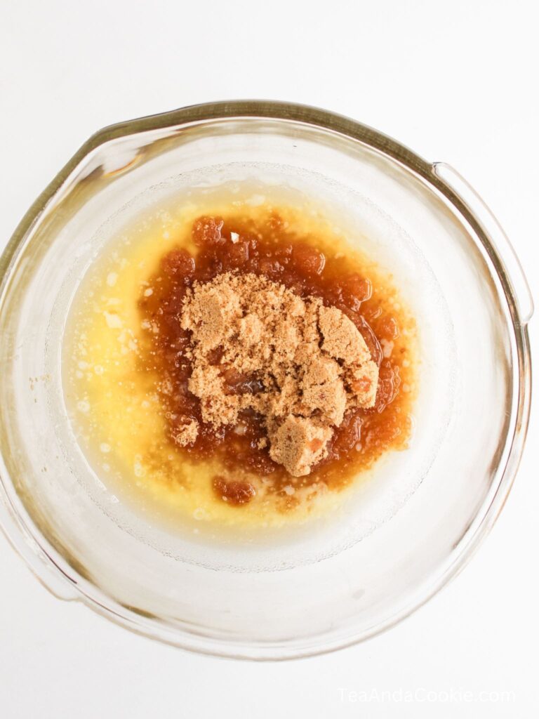 A glass mixing bowl contains melted butter, brown sugar, and what appears to be vanilla extract, partially mixed together.