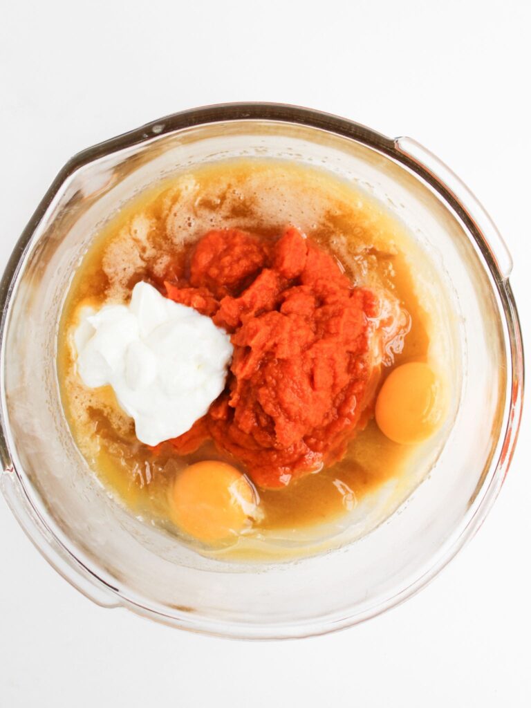 A glass mixing bowl containing pumpkin puree, two raw eggs, yogurt, and a liquid mixture on a white surface.