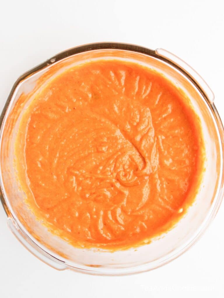 A clear glass bowl filled with smooth, orange-red sauce or batter, viewed from above on a white background.