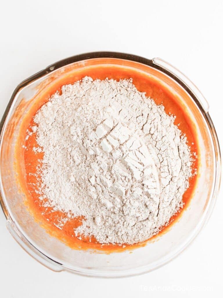A glass bowl with orange batter and a mound of flour on top, ready to be mixed, on a white surface.