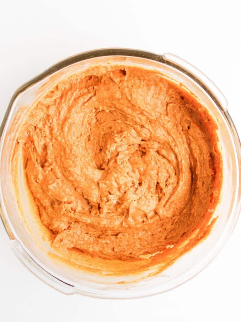 A glass mixing bowl filled with a thick, light brown batter or dough, viewed from above on a white background.