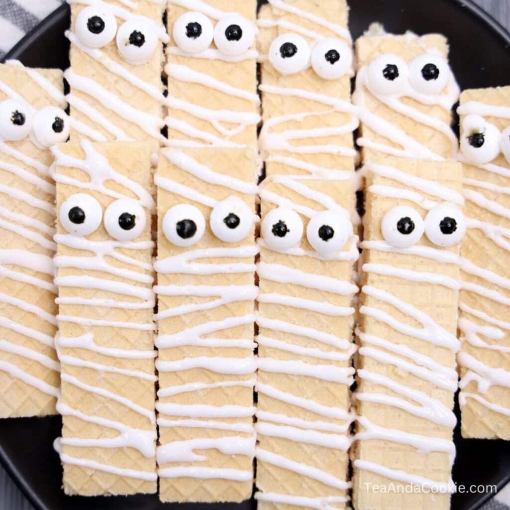 Wafer Mummy Cookies. Rectangular wafer cookies decorated with white icing and candy eyes to resemble mummies, arranged on a black plate.