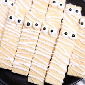 Wafer cookies decorated with white icing and candy eyes to resemble mummies, arranged closely together on a black plate.