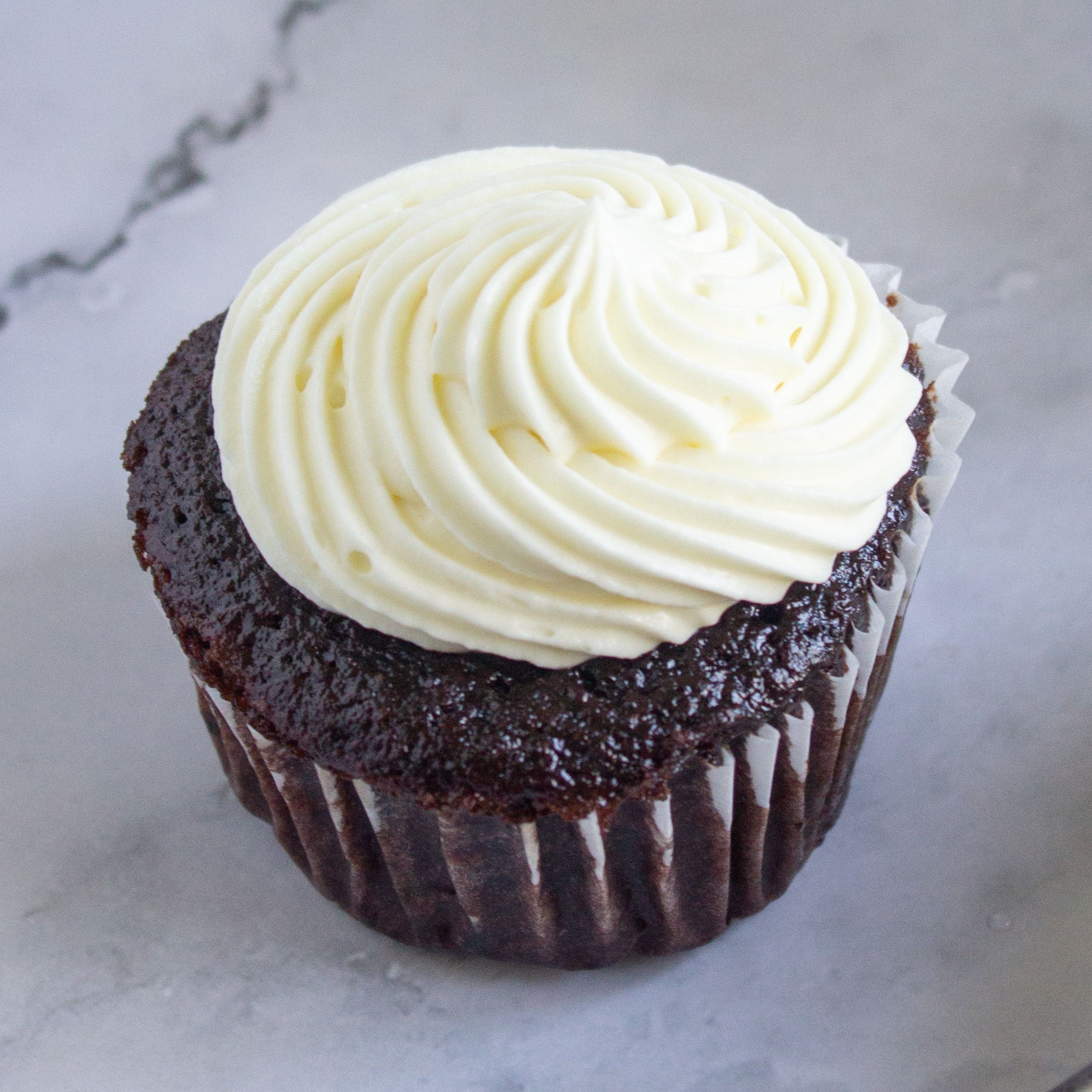 Chocolate cupcakes with sour cream topped with white swirled frosting on a marble surface.