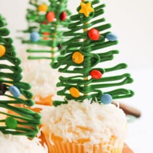 Cupcake topped with white frosting and shredded coconut, decorated with a green chocolate Christmas tree and colorful candy ornaments.
