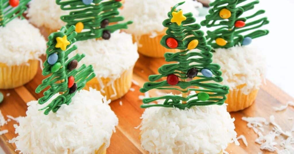 Cupcakes topped with white shredded coconut and decorated with green icing in the shape of Christmas trees, featuring small candy ornaments and stars.