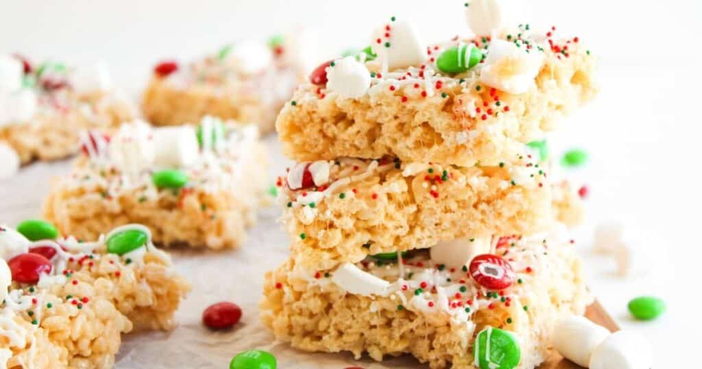 Rice Krispie holiday treats. Three rice crispy treats stacked on top of each other, decorated with white drizzle, mini marshmallows, red and green candy, and sprinkles. More treats and candies are in the background.