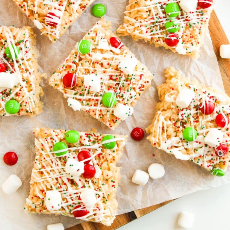 Rice Krispies treats topped with white icing drizzle, red and green M&Ms, sprinkles, and mini marshmallows on parchment paper.