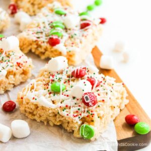 Rice Krispies treats topped with white drizzle, red and green candies, mini marshmallows, and sprinkles on parchment paper.