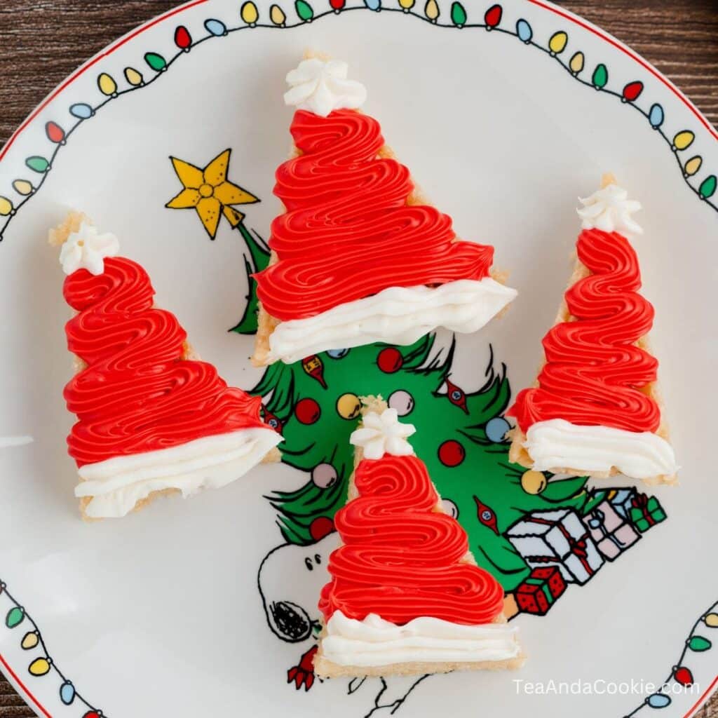 Rice Krispie Santa Hats. Four Christmas tree-shaped cookies with red and white icing are arranged on a festive plate featuring Snoopy and a decorated Christmas tree.