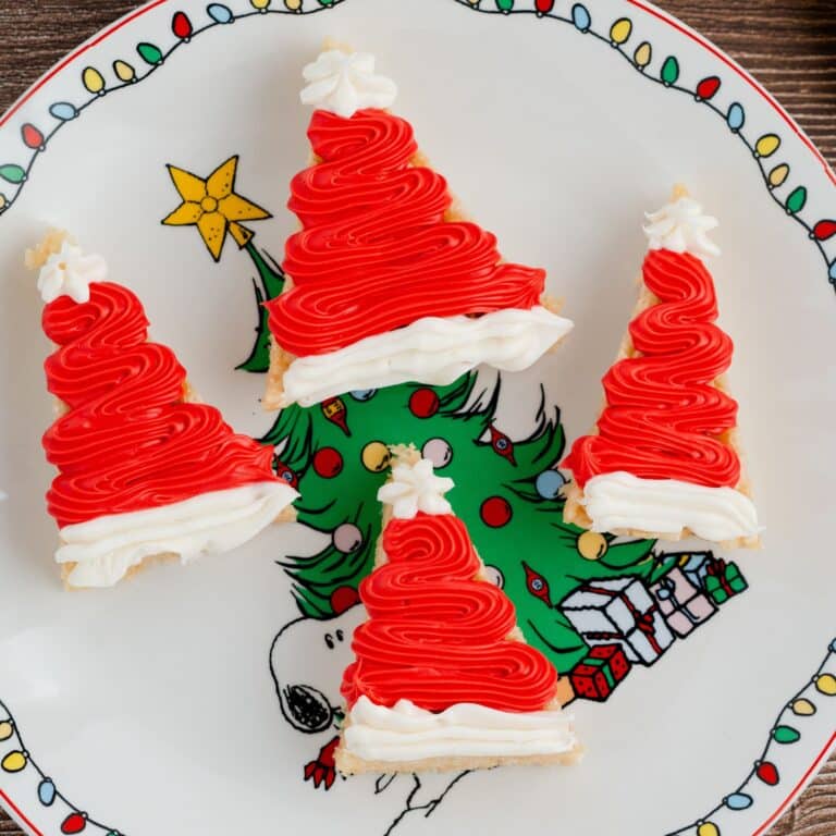 Four triangular cookies decorated to look like Santa hats with red and white icing are arranged on a Christmas-themed plate featuring Snoopy and a Christmas tree.