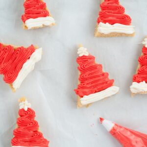 Triangle-shaped cookies decorated with red and white icing to look like Santa hats are arranged on parchment paper, with a piping bag nearby.