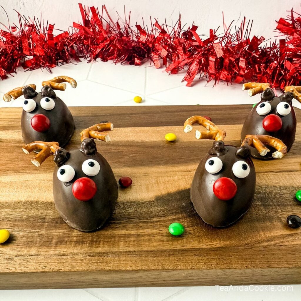 Rudolph Chocolate Covered Marshmallow Candy Recipe. Four chocolate reindeer-shaped treats with pretzel antlers, candy eyes, and red noses are arranged on a wooden board with red tinsel and colorful candies in the background.