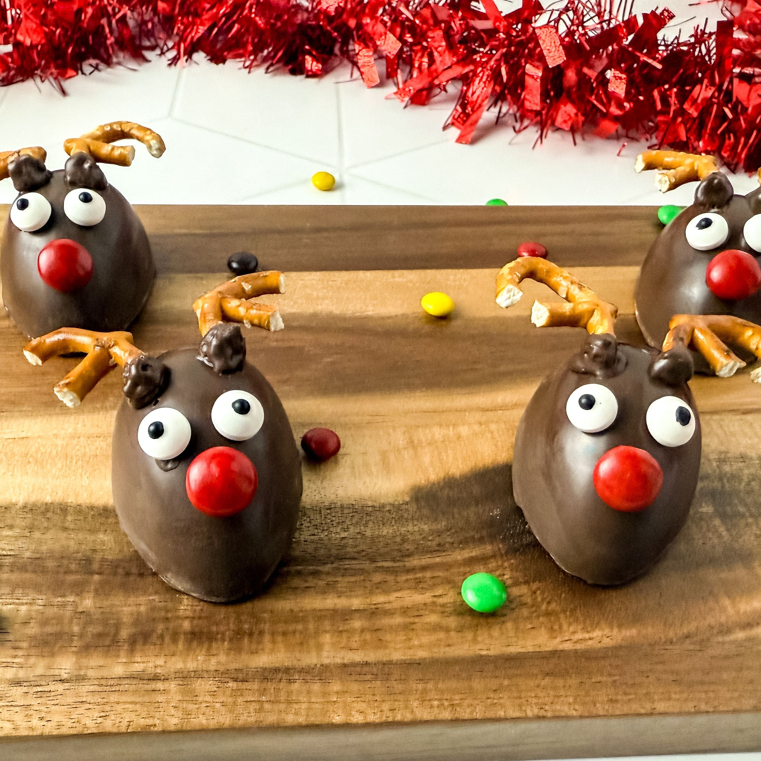 Four reindeer-shaped chocolate treats with pretzel antlers, candy eyes, and red noses are arranged on a wooden board with colorful candy pieces around them.