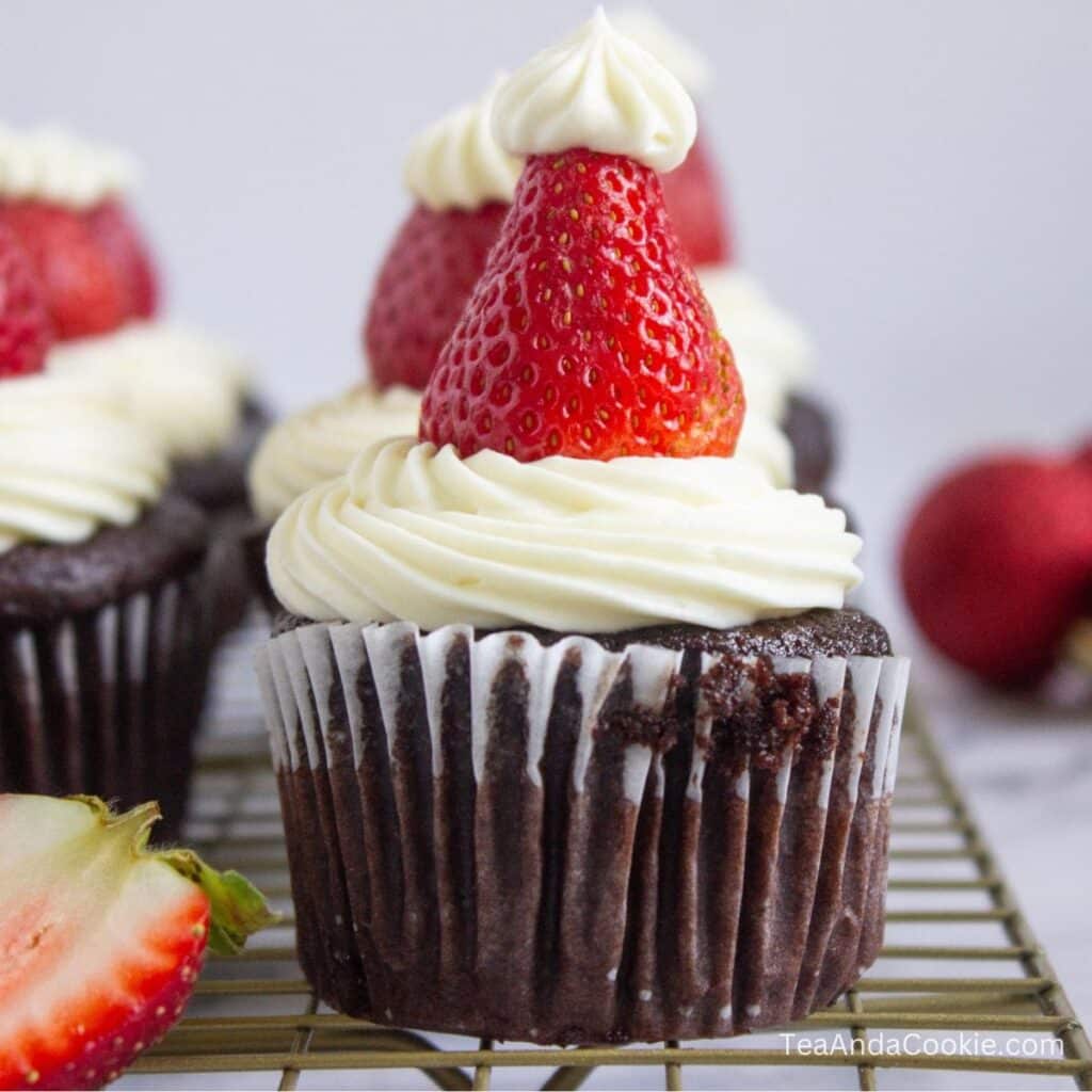 Santa Hat Cupcakes. A chocolate cupcake topped with white frosting and a strawberry, decorated to resemble a Santa hat, sits on a cooling rack.