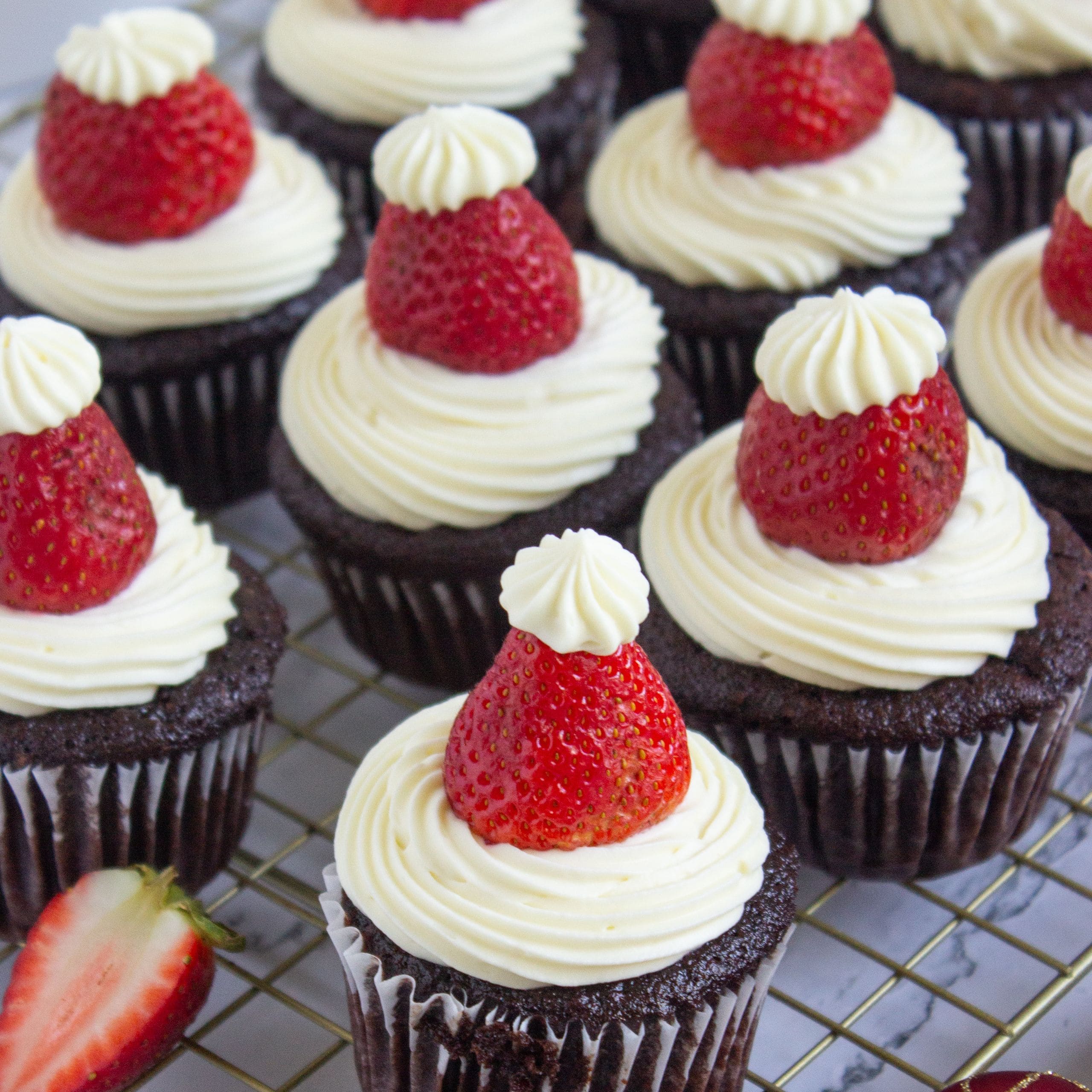Chocolate cupcakes topped with swirls of white frosting and whole strawberries, each with a small dollop of frosting, arranged on a wire cooling rack.