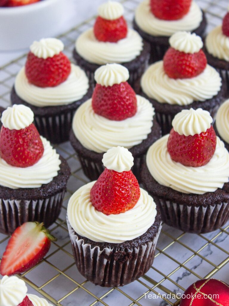 Santa Hat Cupcakes. Chocolate cupcakes topped with swirled white frosting and strawberries designed to look like Santa hats, arranged on a cooling rack.