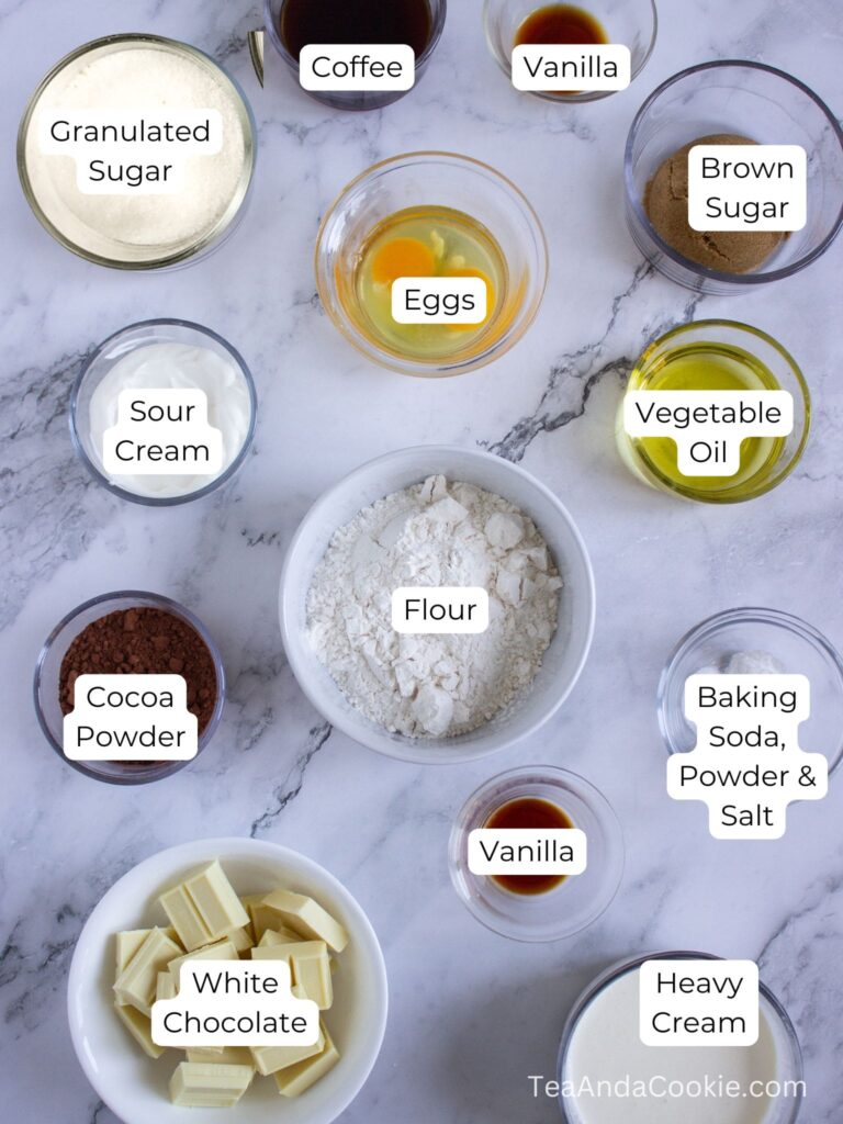 Top-down view of labeled baking ingredients in bowls, including flour, cocoa powder, sugars, eggs, sour cream, oils, white chocolate, heavy cream, coffee, and vanilla.