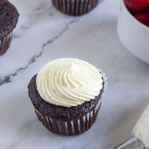 A chocolate cupcake with white swirled frosting sits on a marble surface next to a bowl of strawberries and an icing piping bag.