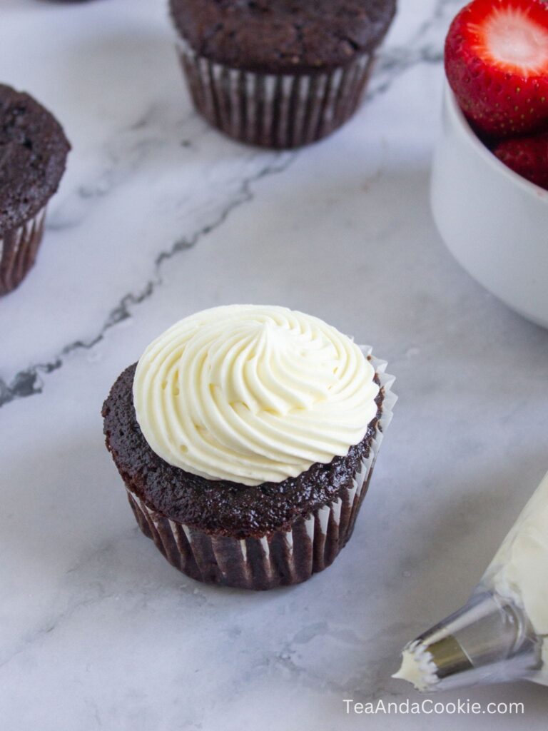 A chocolate cupcake with white swirled frosting sits on a marble surface next to a bowl of strawberries and an icing piping bag.