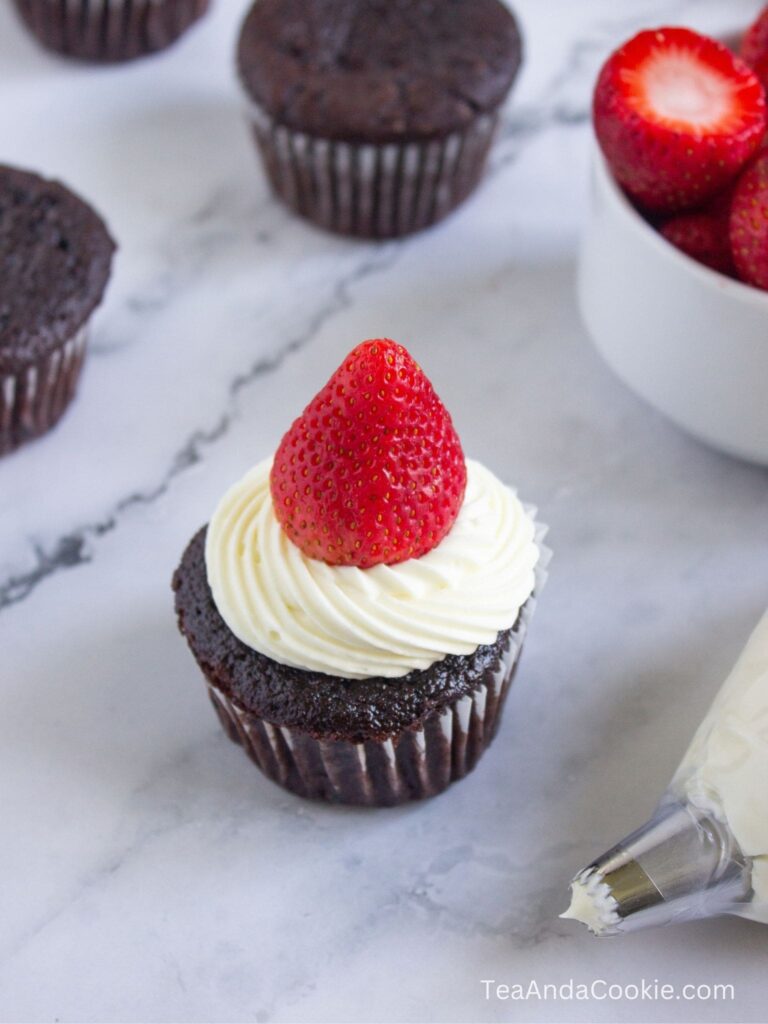 A chocolate cupcake with white frosting and a whole strawberry on top sits on a marble surface, with other cupcakes and a bowl of strawberries in the background.