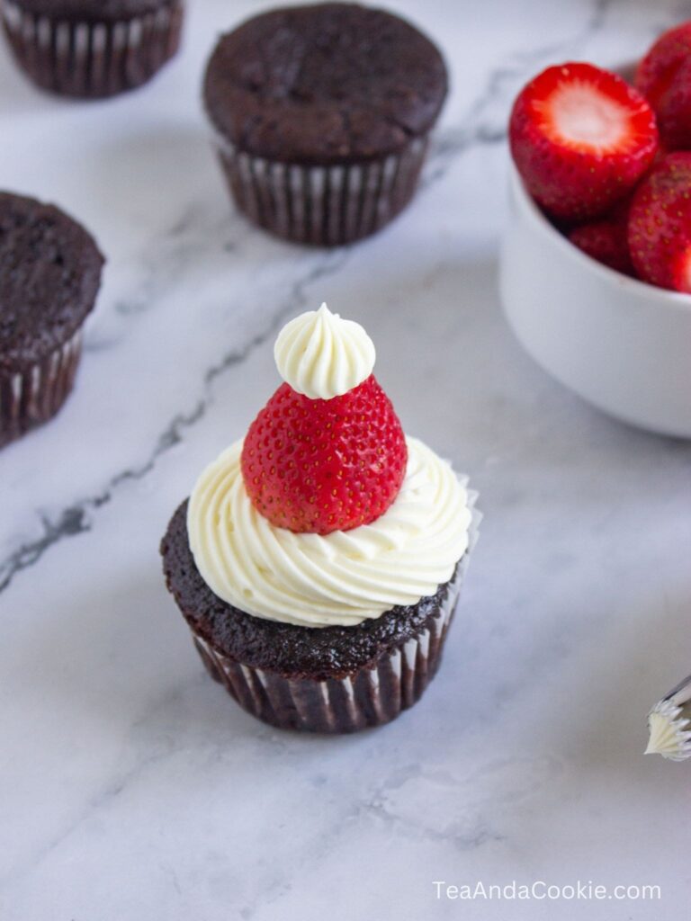 A chocolate cupcake with white frosting, topped with a whole strawberry and a small dollop of frosting, sits on a marble surface near other cupcakes and a bowl of strawberries.