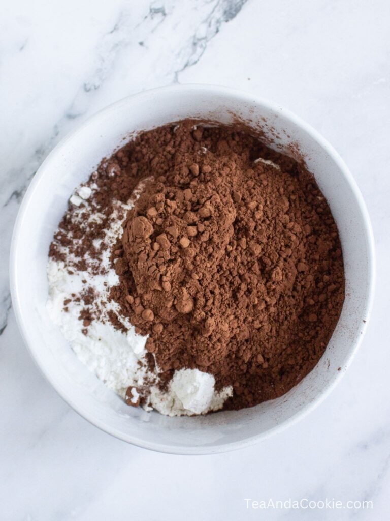 A white bowl containing unsweetened cocoa powder and flour sits on a marble countertop.