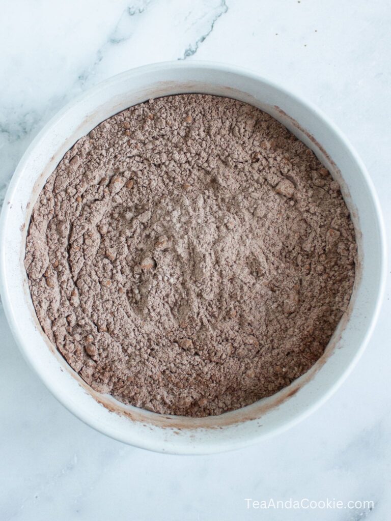A white bowl filled with sifted cocoa powder and dry baking ingredients on a marble surface.