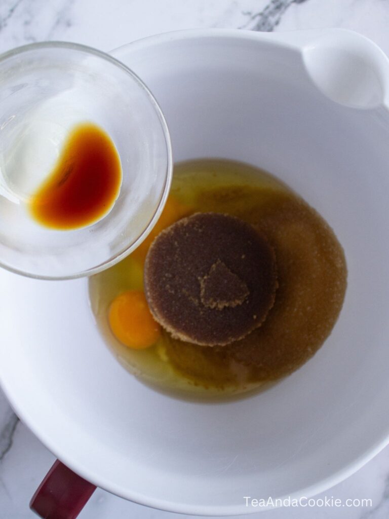 A mixing bowl with eggs, brown sugar, and granulated sugar, while a small cup of vanilla extract is being poured in.