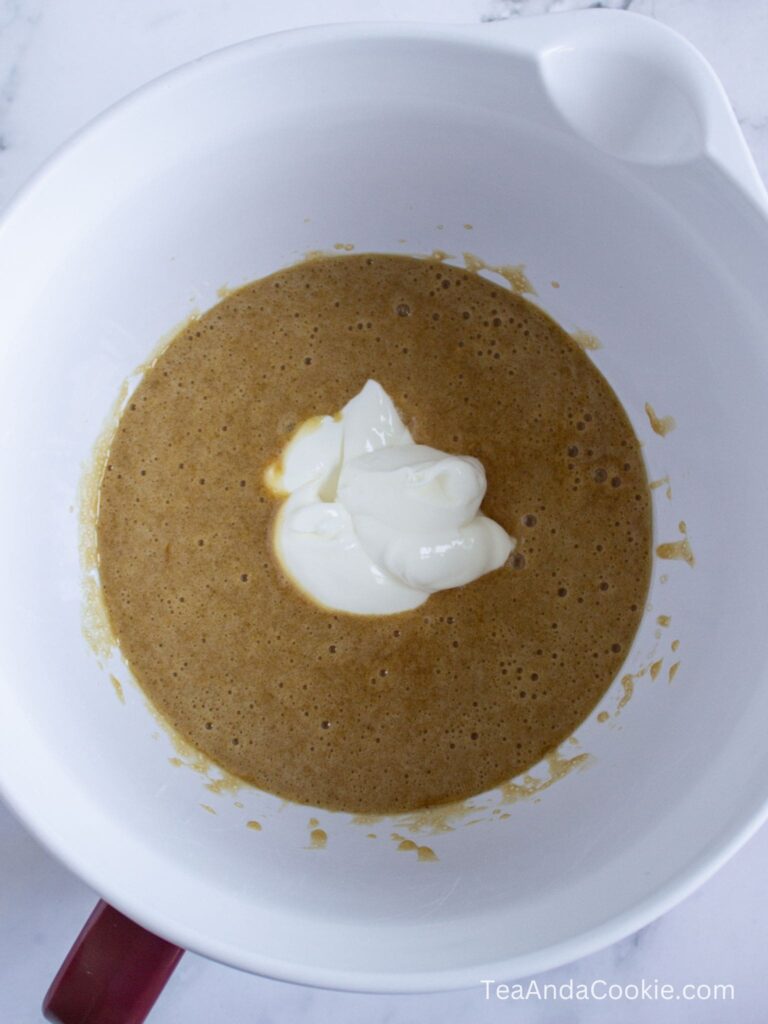 A white mixing bowl with brown batter and a dollop of white yogurt or sour cream in the center, sitting on a marble countertop.