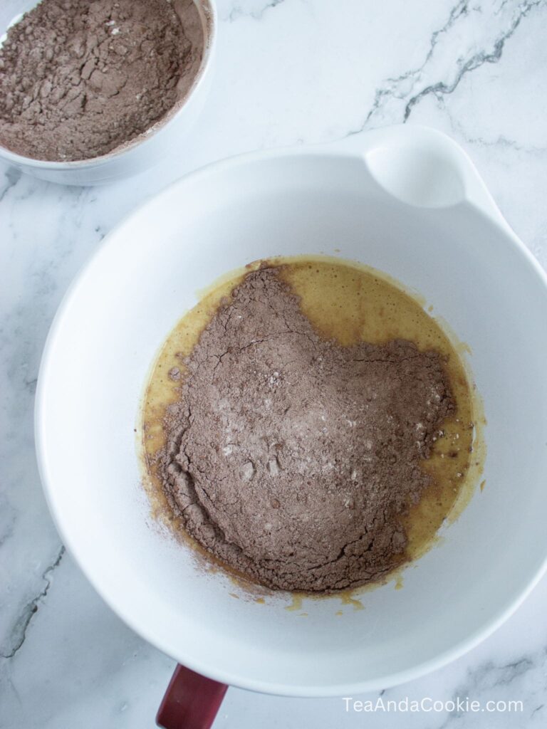 A white mixing bowl with eggs and cocoa powder mixture, with another bowl of dry cocoa mix in the background on a marble countertop.