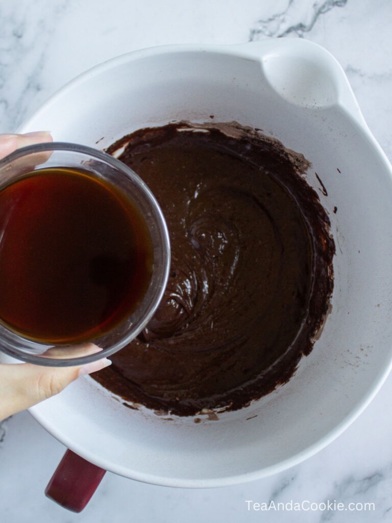 A hand holds a glass bowl of liquid above a mixing bowl filled with chocolate batter on a marble surface.