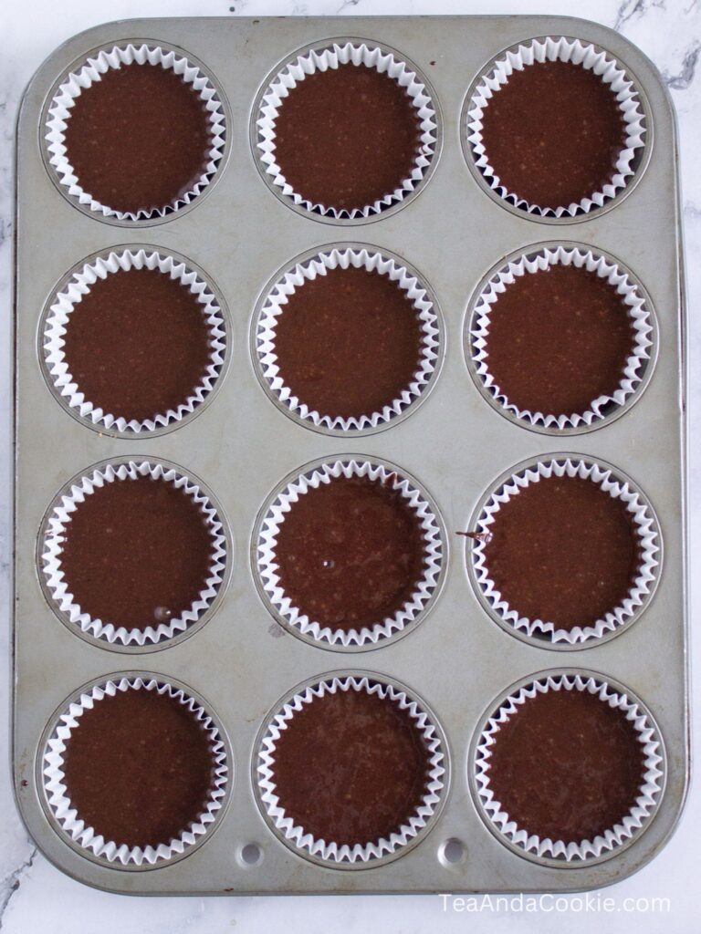A muffin tin filled with twelve cupcake liners, each containing chocolate batter, ready to be baked.