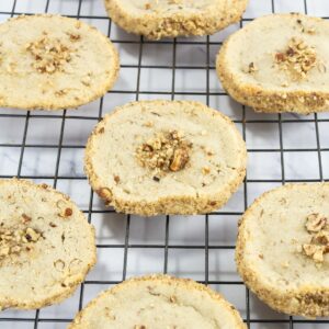 Round shortbread cookies with chopped nuts on top and around the edges, cooling on a black wire rack over a white marble surface.