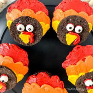Cupcakes decorated with red, orange, and yellow frosting to resemble turkeys, featuring cookie faces with candy eyes and frosting beaks, arranged on a black plate.